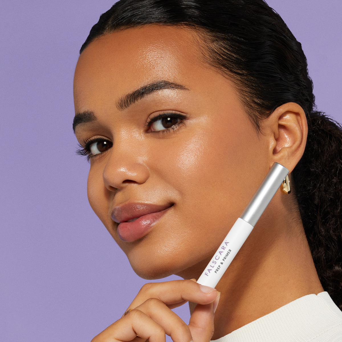 A woman holds a mascara tube labeled &quot;Falscara&quot; near her face, with a smooth complexion and natural makeup, against a purple background.