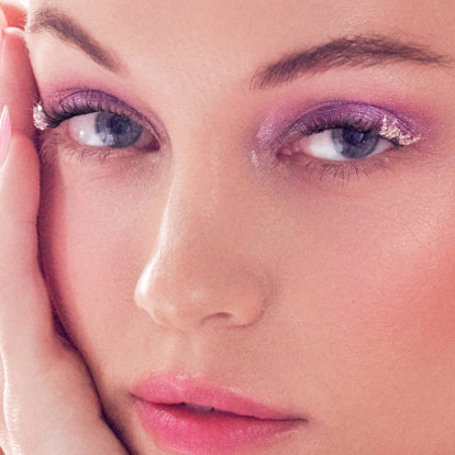 Close-up of model's face with purple eyeshadow, pink lips, and hand near face.