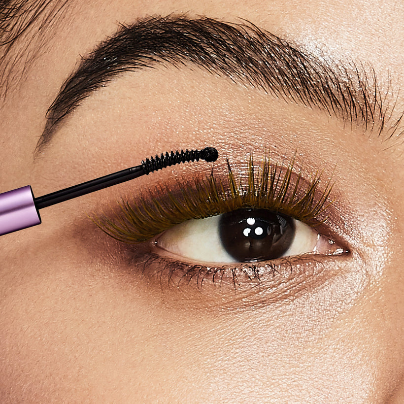 Mascara wand applying black mascara to upper lashes with gold-toned eyeshadow.
