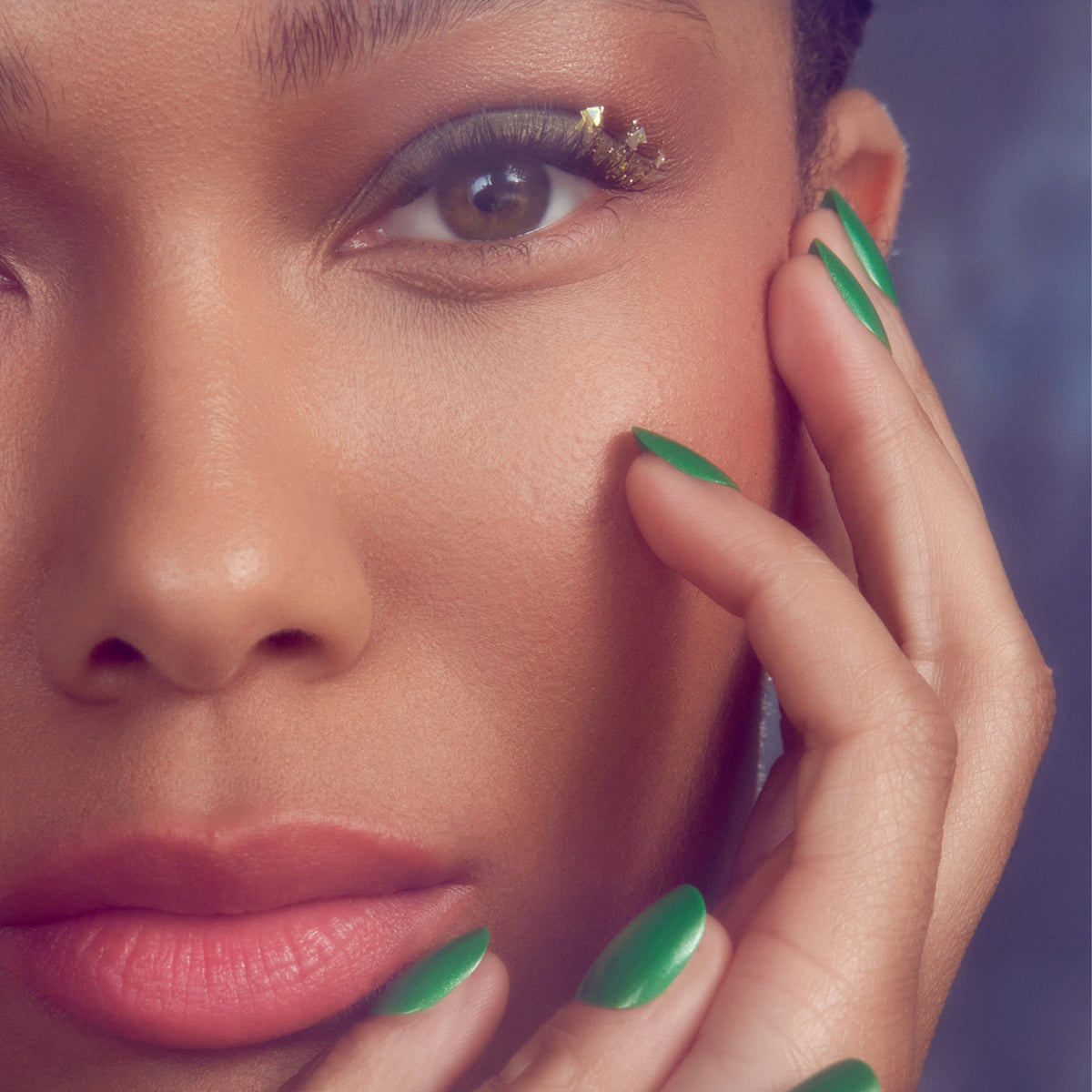 Woman with gold eyeshadow and green nail art touching face against purple background.