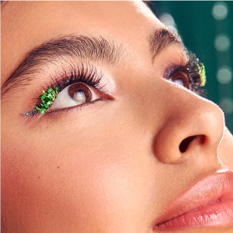 Close-up of eye makeup featuring green glitter eyeliner and black mascara against blurred background.
