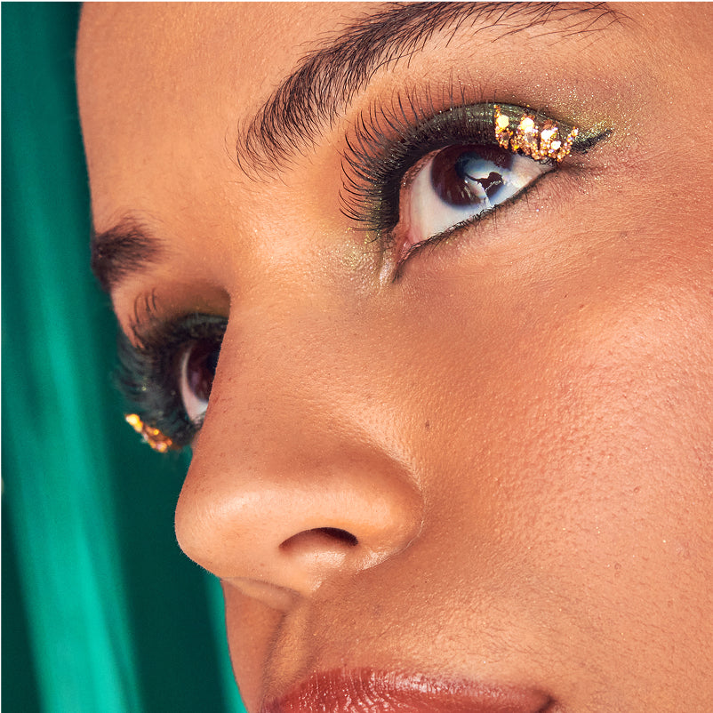 Close-up of eye makeup featuring gold glitter eyeliner and false lashes on model against teal background.